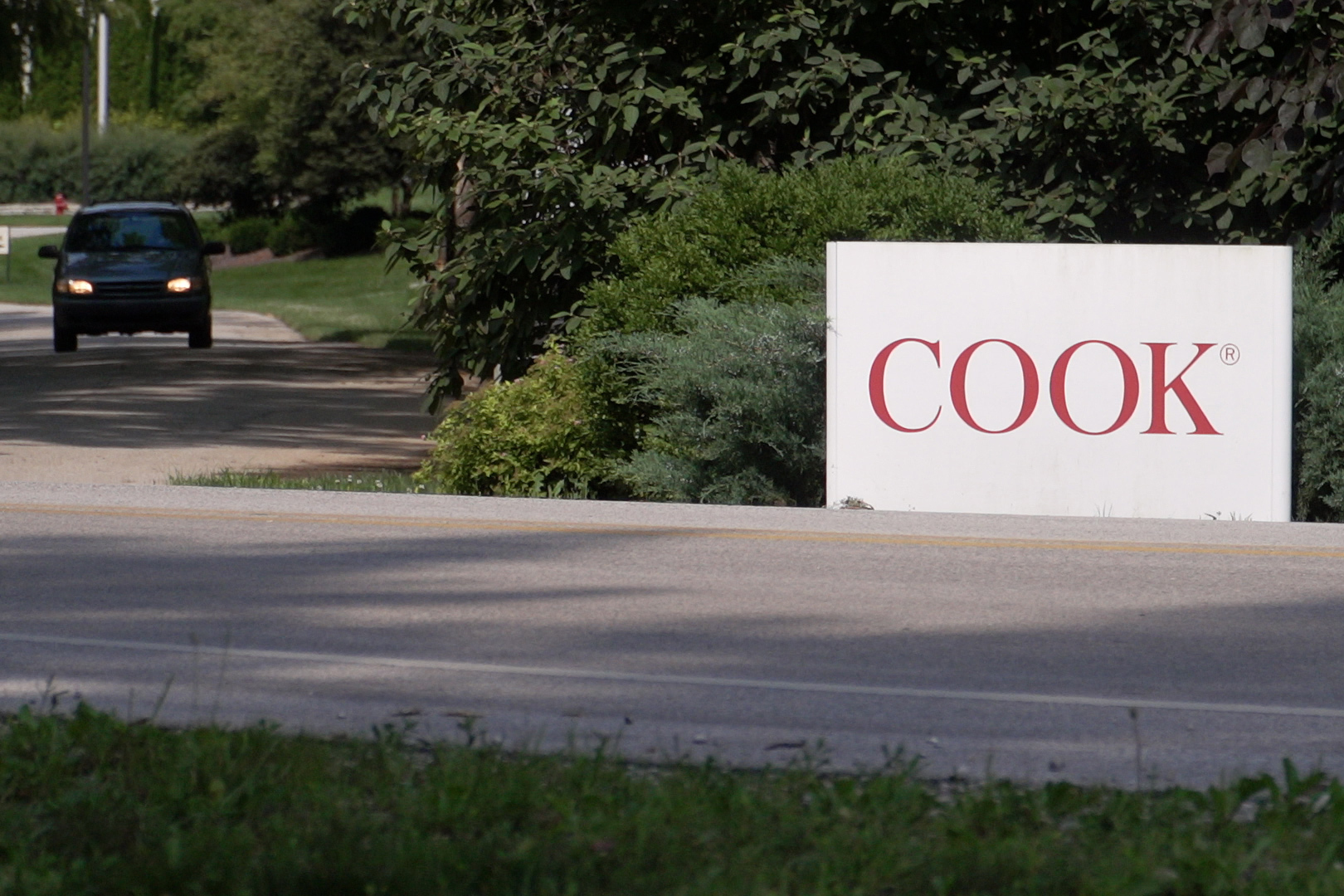 The entrance to Cook Group Inc. in Ellettsville. A clean white sign sits on the side of the road. Medium red letters spell out Cook in all caps. To the left of the sign, a mid-sized vehicle with its lights on drives up a path. Behind the sign, green foliage. Image credit: Alan Mbathi, IPB News