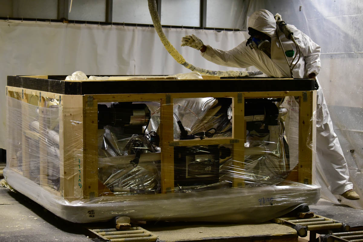 A worker in a fully enclosed protective suit sprays an insulating foam around the components of a nearly completed spa. Image credit: Justin Hicks, IPB News