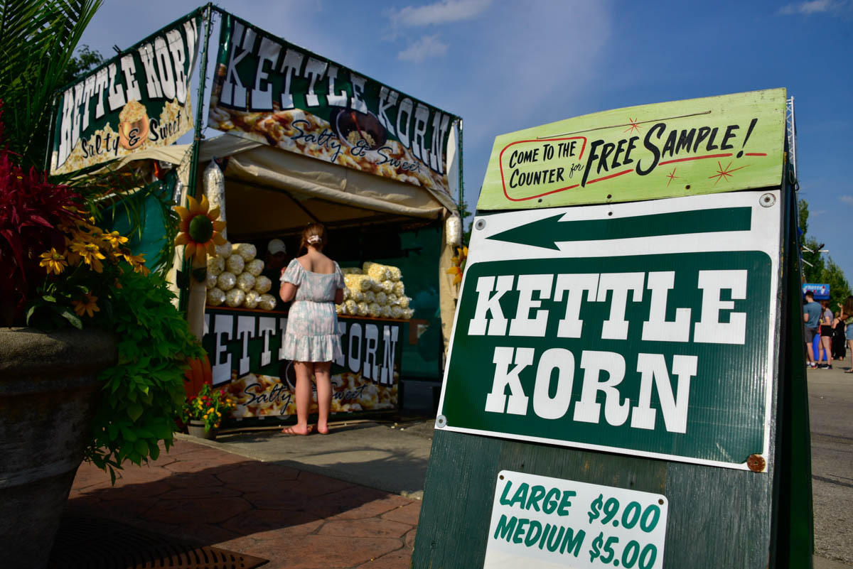 A customer orders a bag of kettle corn at Rairigh's booth during the Indiana State Fair. A large green sign in the foreground reads KETTLE KORN and promises free samples at the counter. Image credit: Justin Hicks, IPB News
