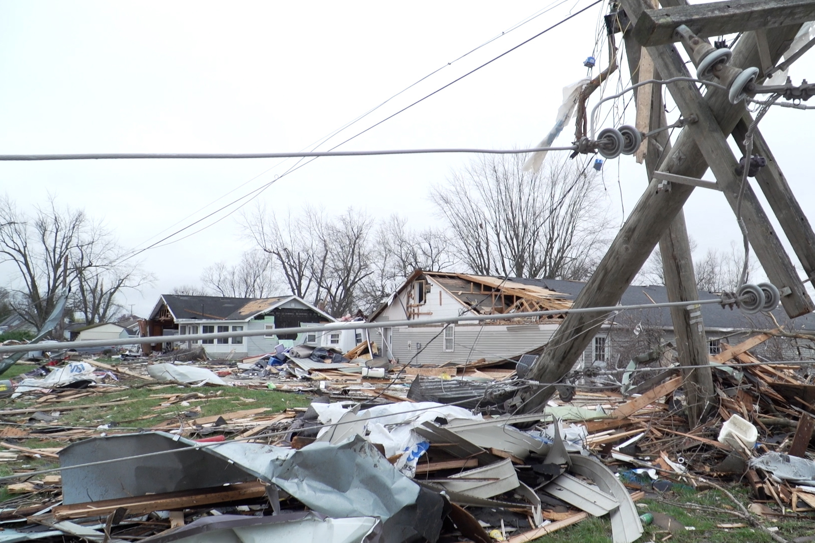 A power line is down with cables partially obstructing the view of demolished and damaged homes. Debris is scattered across grass and stacked by crews.