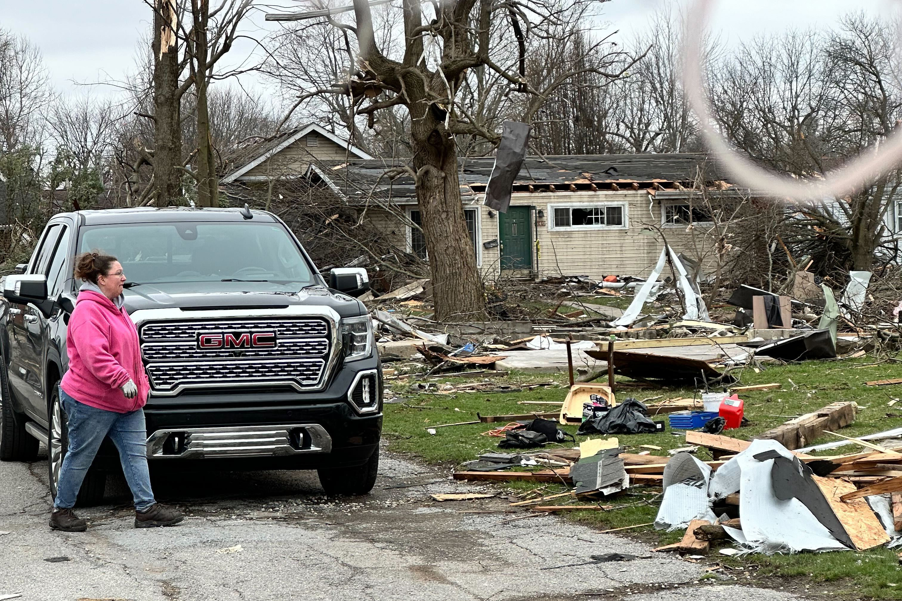 A woman in a pink jacket and gloves walks in front of a pickup truck toward a yard scattered with debris and branches. The home in the background is missing chunks of its roof.