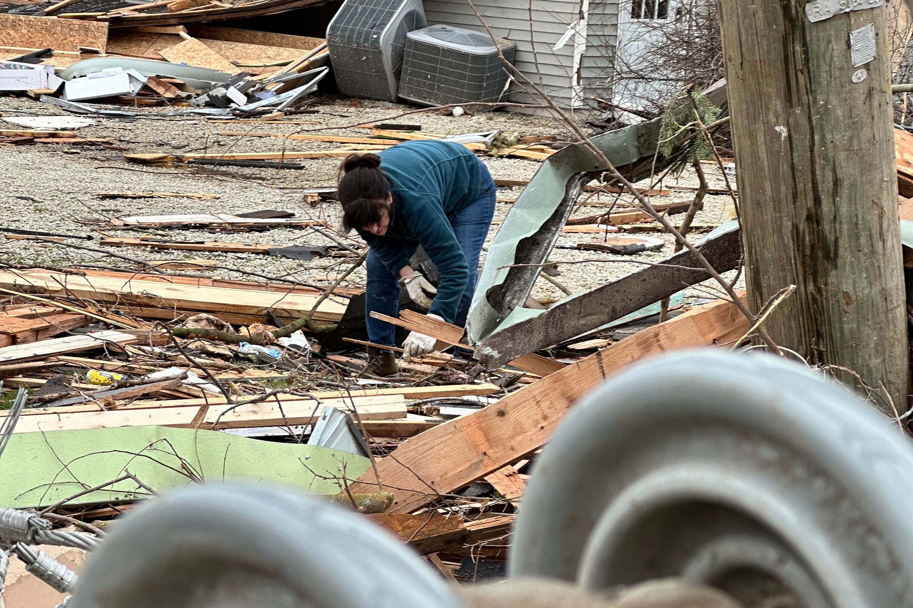 a woman in a green sweater is bent over, picking through debris surrounding her. Beams from the nearby homes are scattered across gravel and grass.