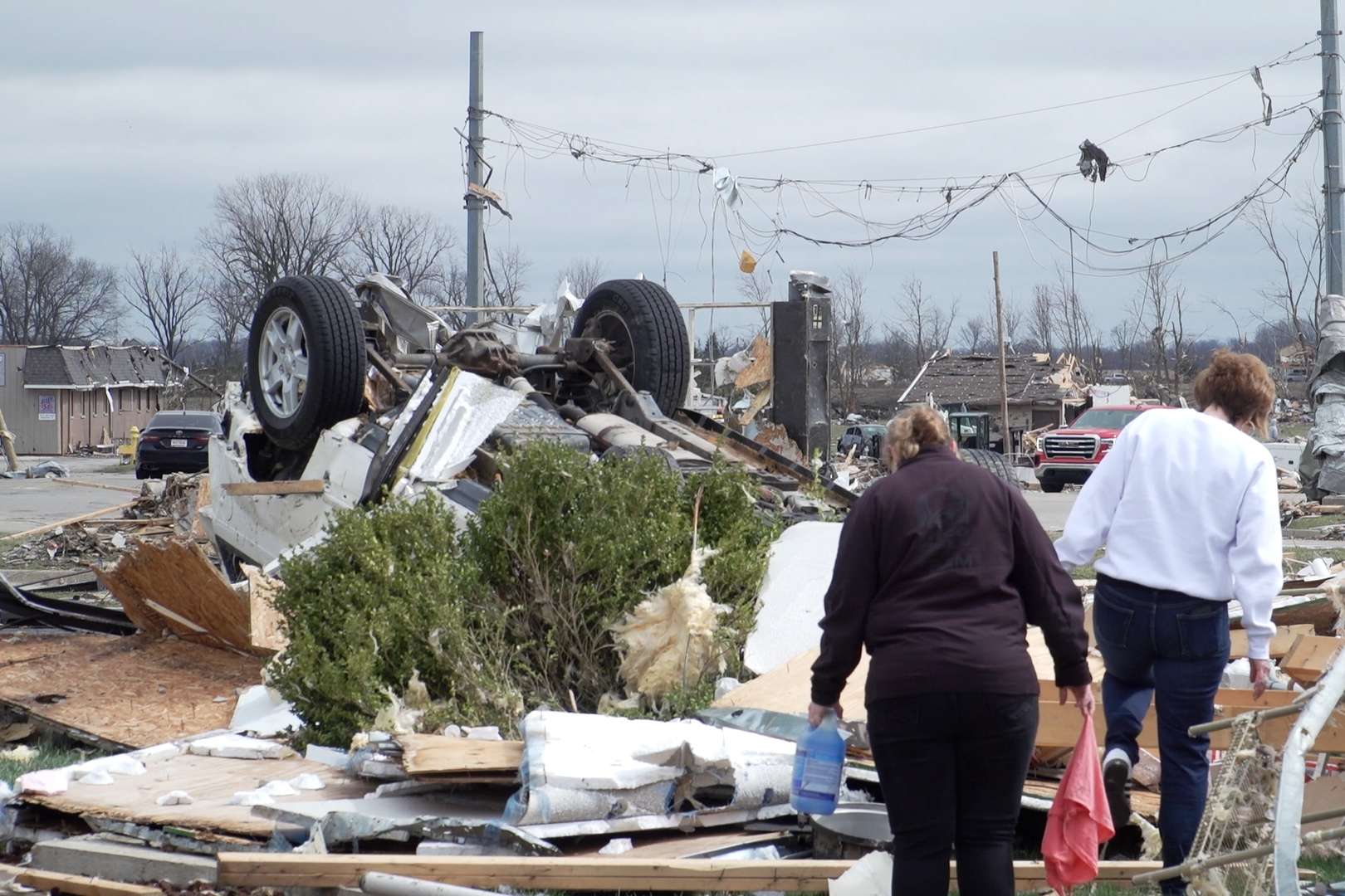 Two people carefully make their way through the debris of the former Taco Bell. The powerlines above them are littered with cords and trash tangled in the wires by the tornado.